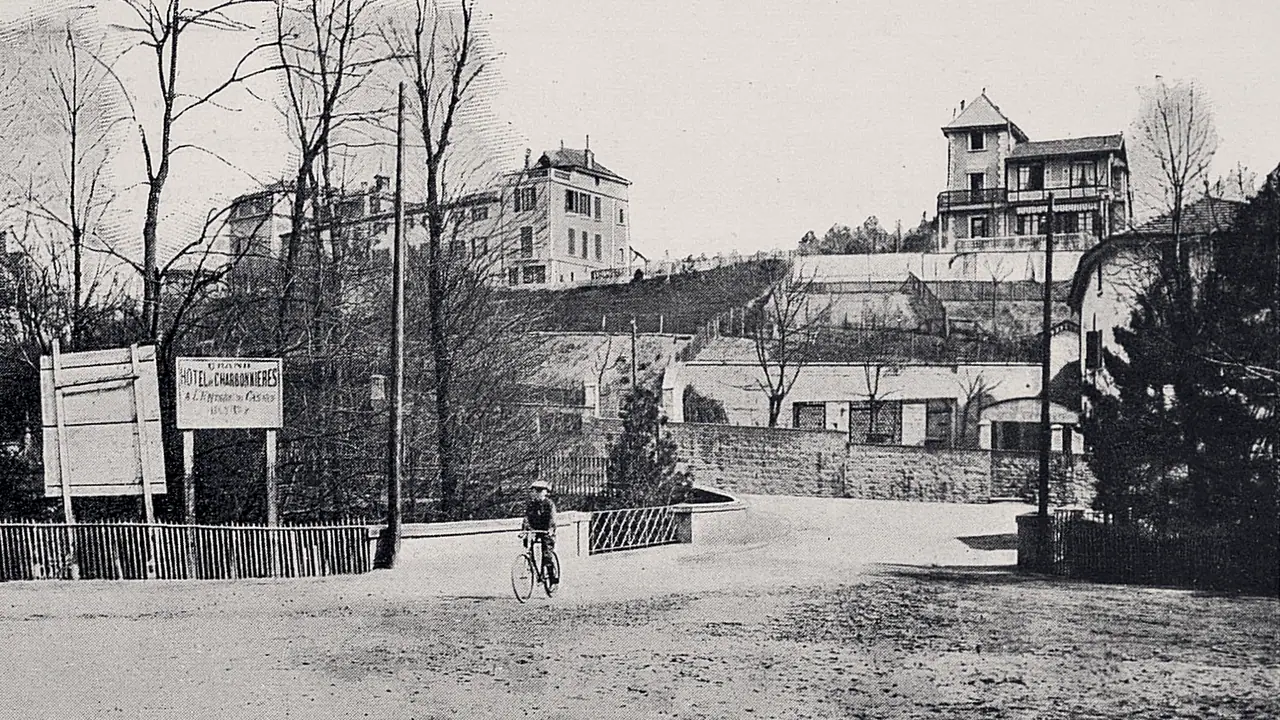 Photographie des années 1900 : une place de terre battue où passe un cycliste, un panneau d'hôtel sur une palissade, et des villas à balcons étagées sur le coteau.