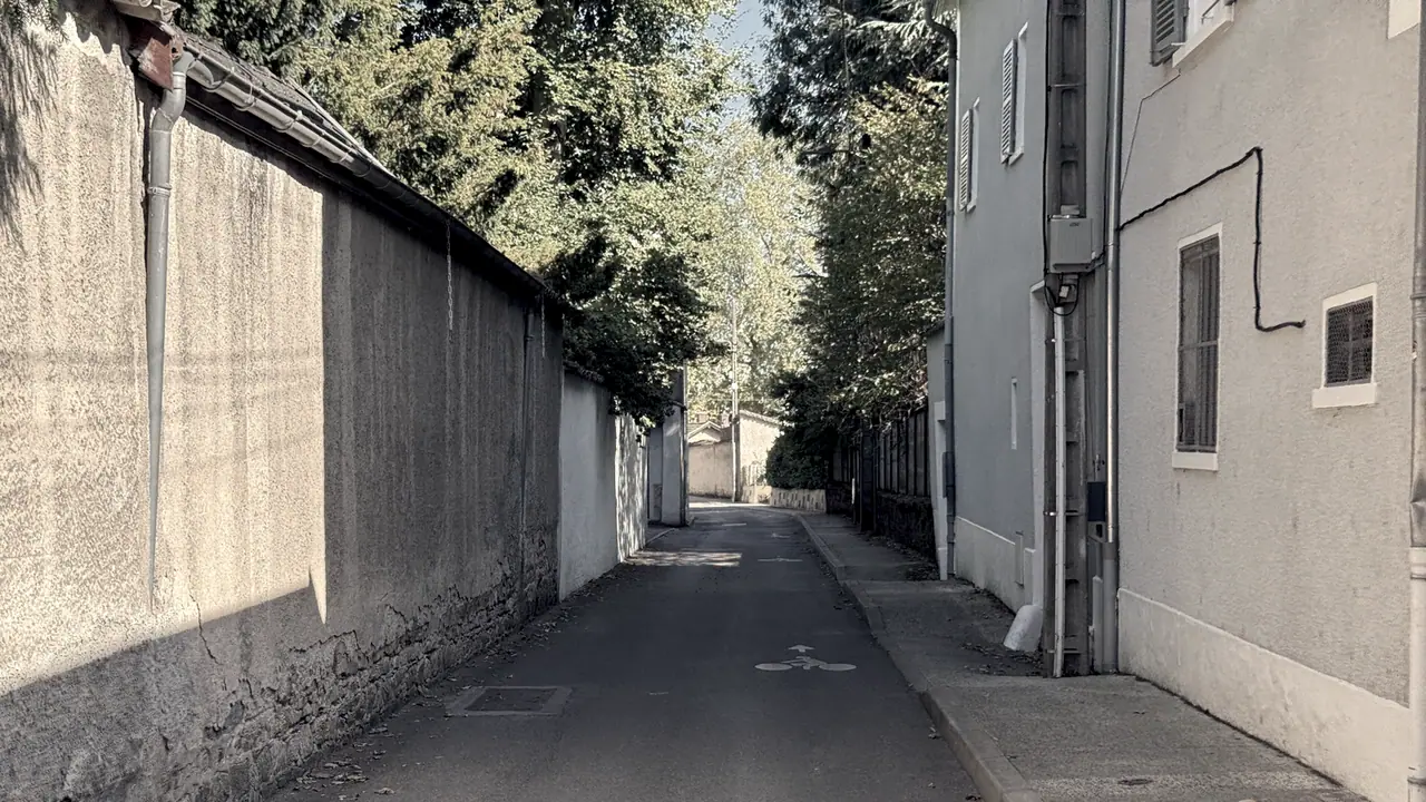 Chemin étroit entre un haut mur crépi aux pierres apparentes et des maisons aux volets gris, avec une bande cyclable peinte au sol et des arbres au fond.