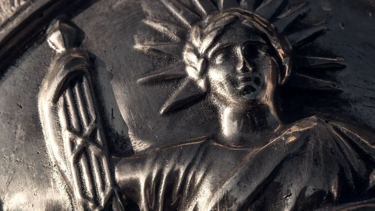Gros plan sur un panonceau de notaire en relief : une figure casquée aux rayons en couronne tient un faisceau, dans la lumière rasante.