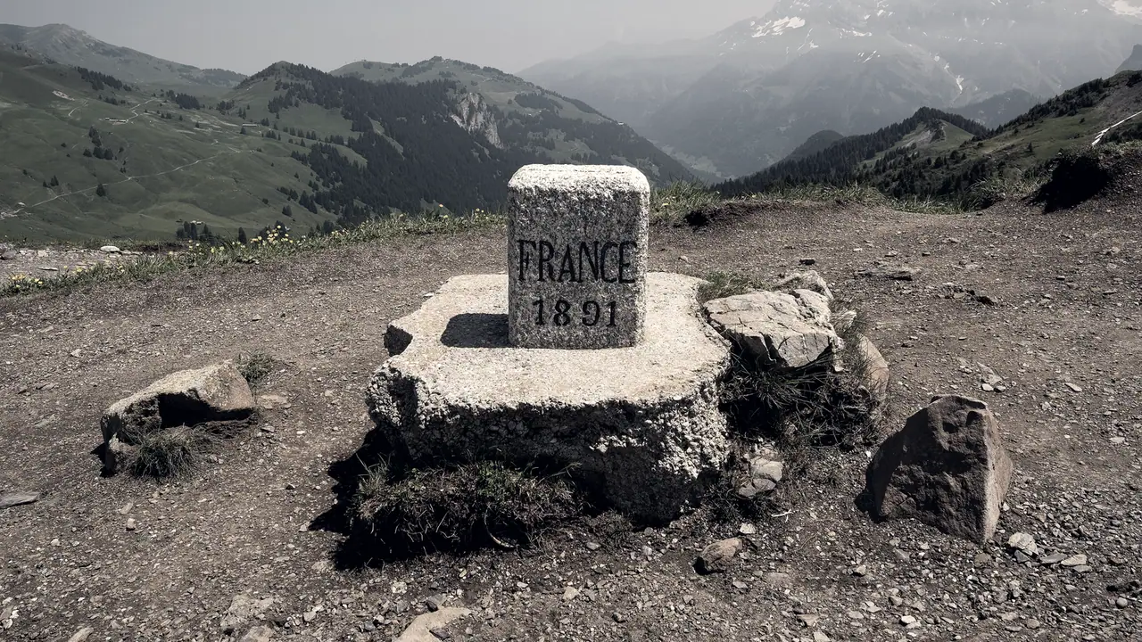 Borne de granit gravée « France 1891 » posée sur un socle de pierre au col de Cou, devant des alpages et un massif enneigé.