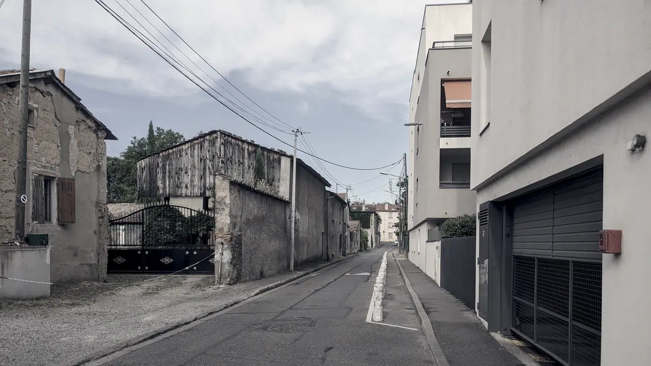 Rue étroite bordée à gauche par une maison ancienne en pierre à volets et un portail en fer forgé, face à un immeuble récent avec porte de garage.
