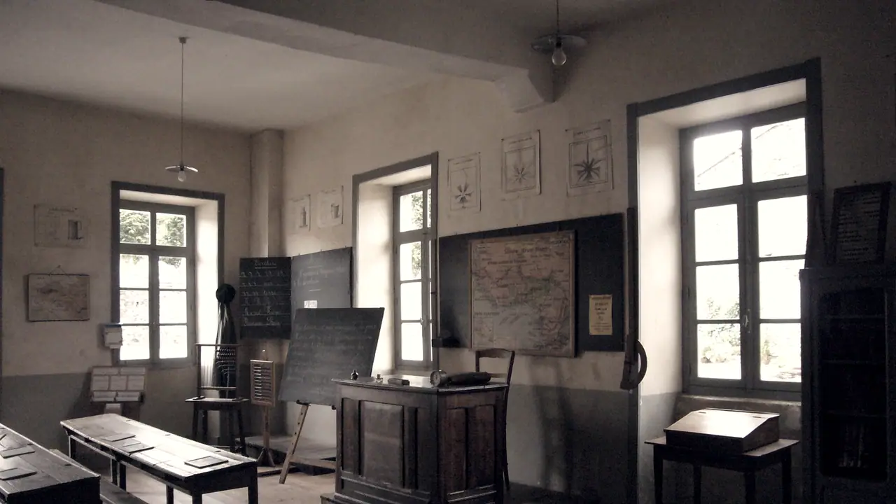 Ancienne salle de classe d'école communale : pupitres de bois au premier plan, bureau du maître au centre, tableaux noirs et carte au mur.