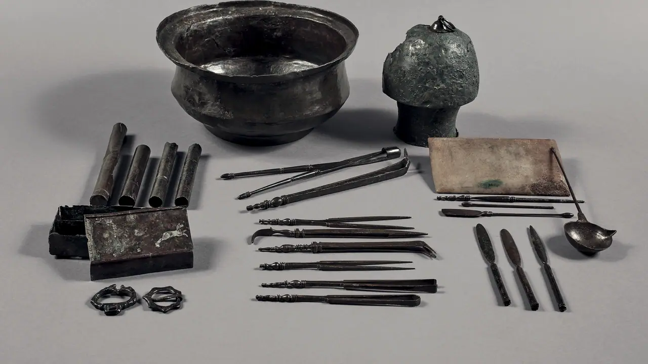 Trousse d'un chirurgien gallo-romain étalée sur un fond gris : un bassin de bronze, des étuis, des pinces et des sondes alignés côte à côte.