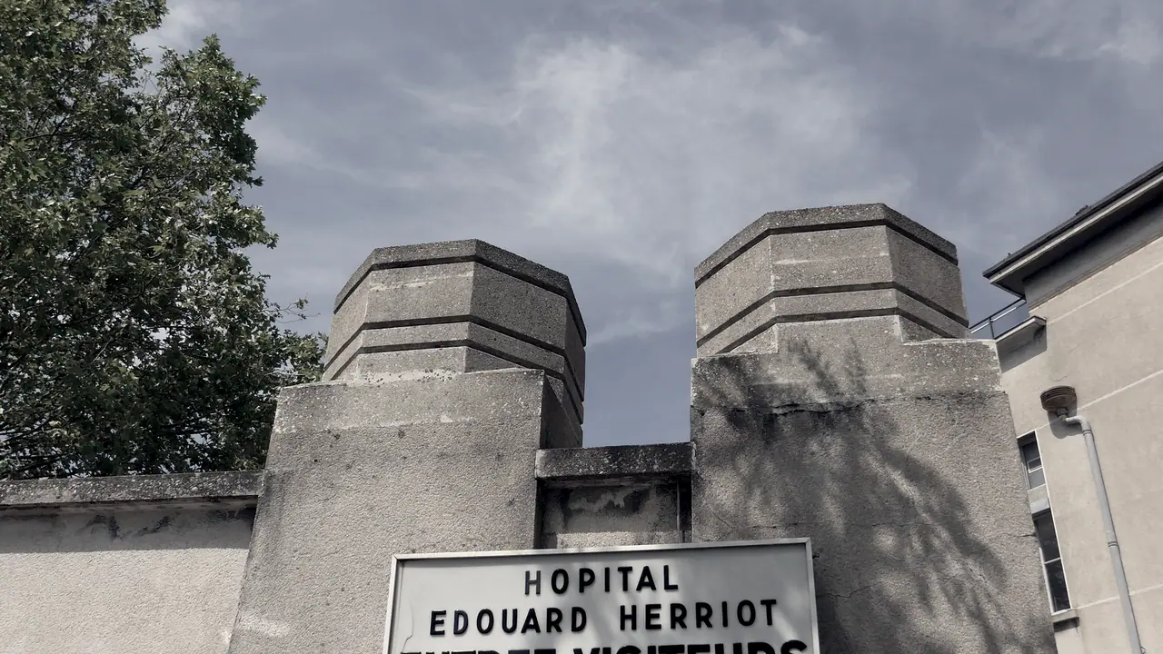 Deux piliers massifs en pierre encadrent l'entrée d'un hôpital, panneau émaillé au bas du cadre sous un ciel nuageux.