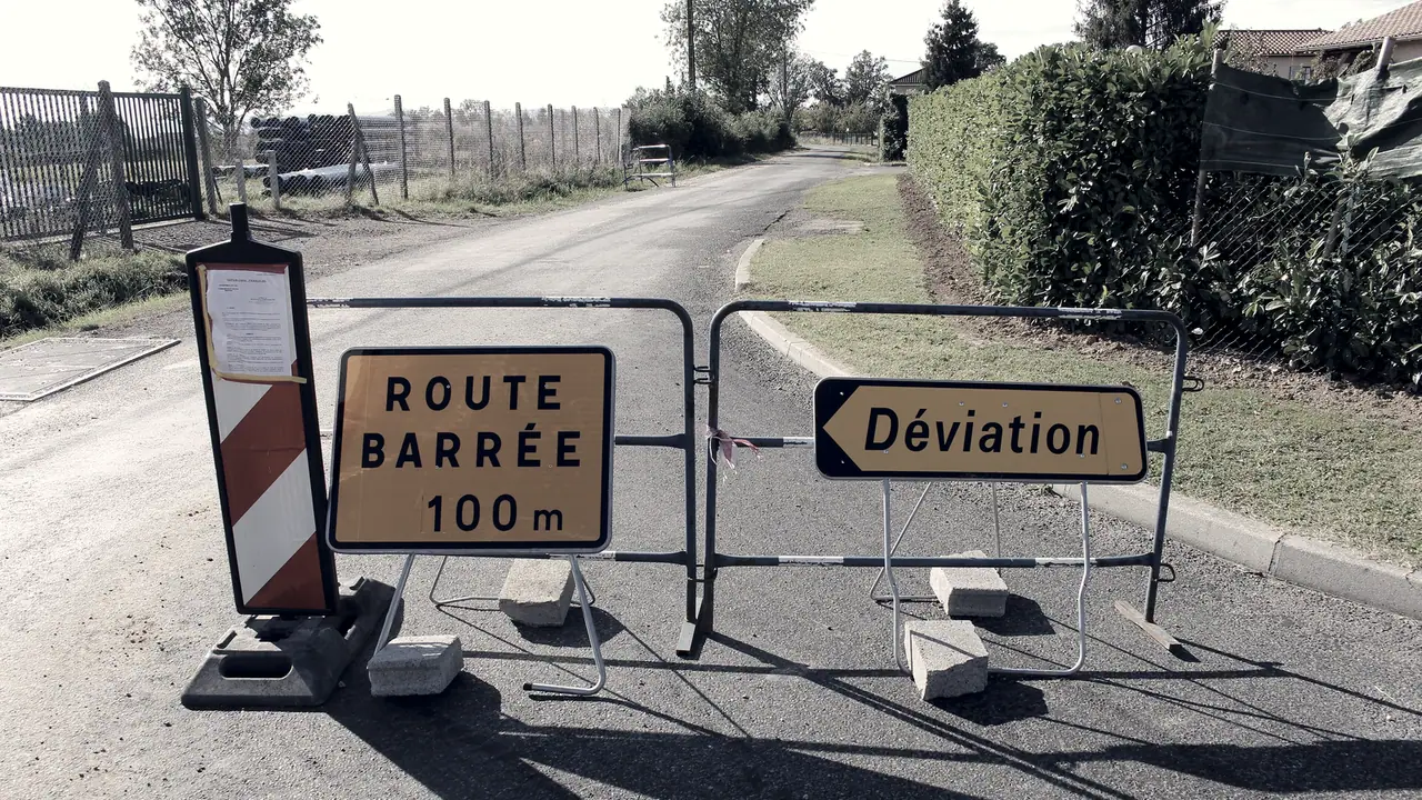 Barrières de chantier en travers d'une petite route, avec un panneau Route barrée à 100 m et une flèche Déviation qui indique un autre chemin.