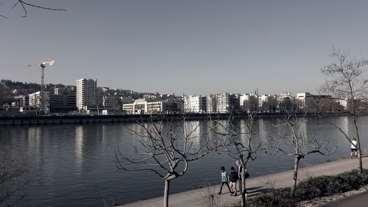 Promeneurs marchant le long d'un chemin au bord d'un fleuve, arbres nus au premier plan et immeubles avec une grue sur l'autre rive.