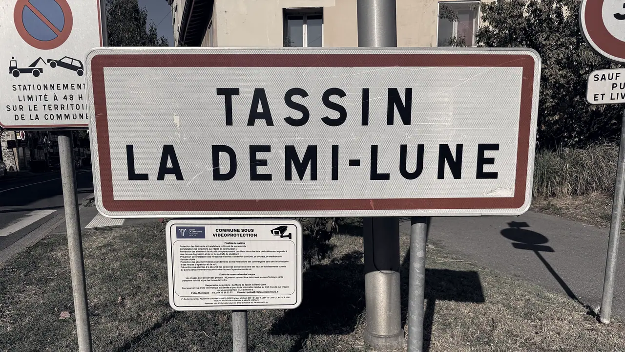 Panneau d'entrée d'agglomération au nom de Tassin-la-Demi-Lune, bordé de rouge, planté dans l'herbe au bord d'une avenue, sous un soleil rasant.