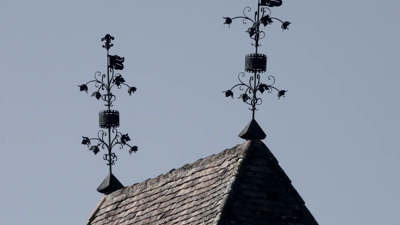 Deux girouettes de fer forgé à fanion et rinceaux plantées au sommet d'un toit de tuiles plates, se découpant sur un ciel uni.