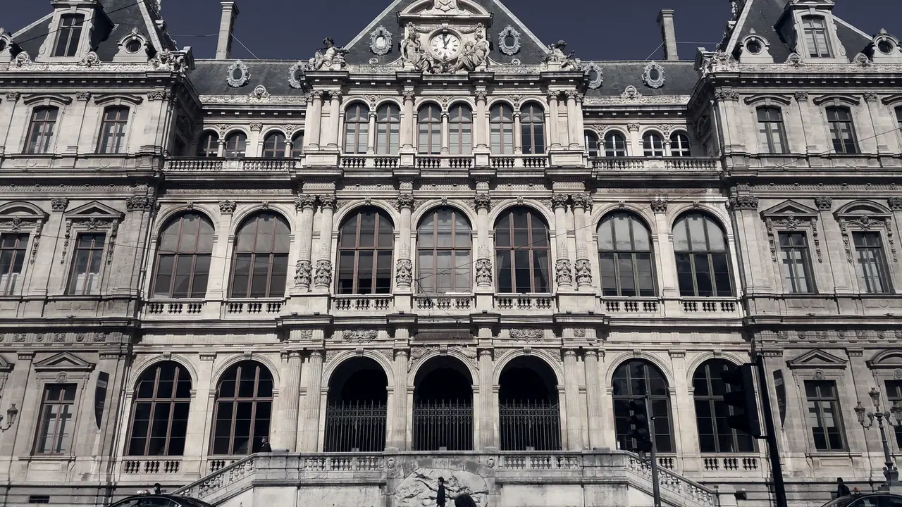 Façade sculptée d'un palais du XIXe siècle avec arcades, colonnes et horloge sous le fronton, escalier de pierre au pied du bâtiment.