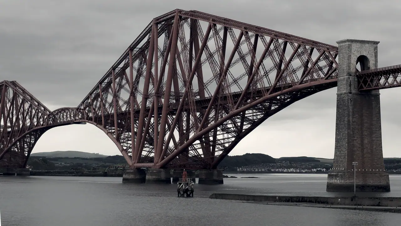 Bras en porte-à-faux d'un pont ferroviaire en treillis d'acier, lancé au-dessus de l'estuaire depuis ses piles de pierre, sous un ciel couvert.