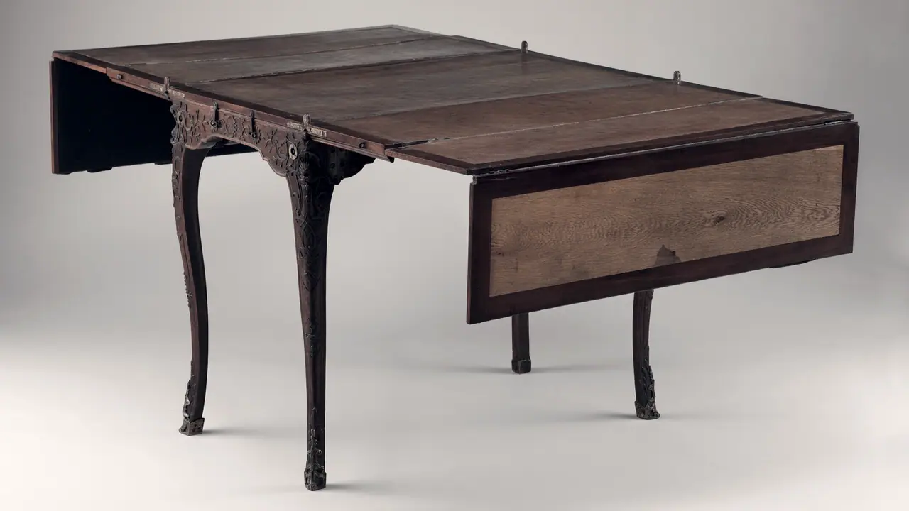 Table de voyage ancienne aux pieds galbés, son plateau de bois prolongé au-delà de la ceinture sculptée et porté par une seconde paire de pieds, sur fond gris uni.