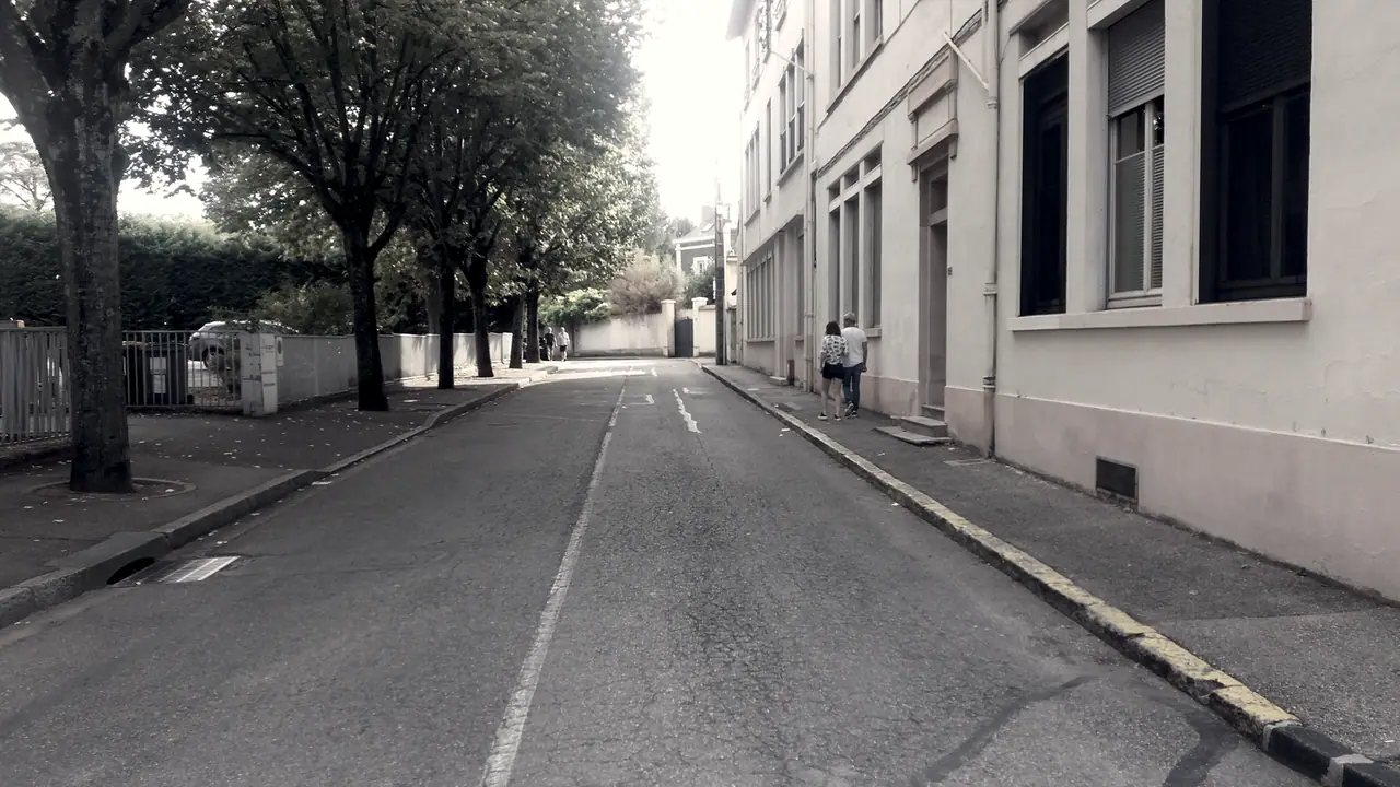 Rue résidentielle déserte vue au ras de la chaussée, arbres et murs de clôture à gauche, deux passants marchant devant les maisons.