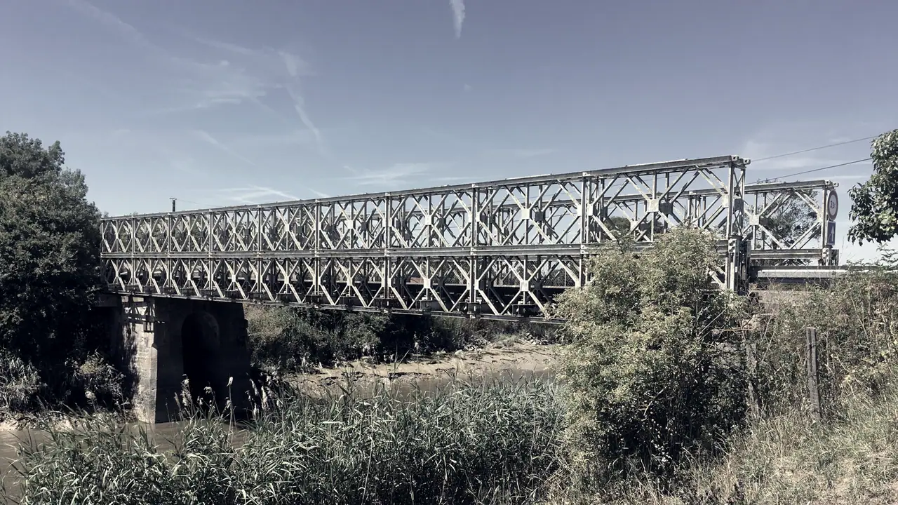 Pont provisoire à poutres d'acier en treillis posé sur une ancienne pile de pierre, au-dessus d'un cours d'eau envahi de roseaux et de broussailles.