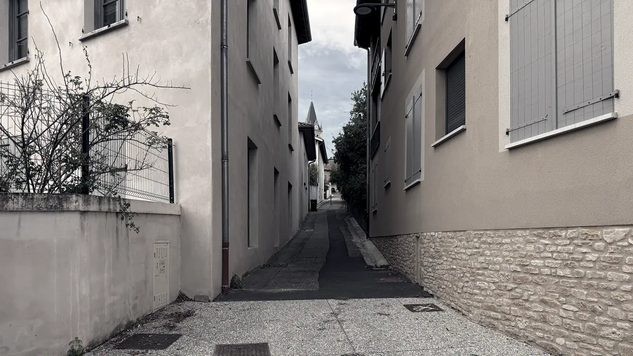 Ruelle étroite entre des maisons à volets, mur en pierre appareillée sur la droite et clocher pointu au bout du passage.