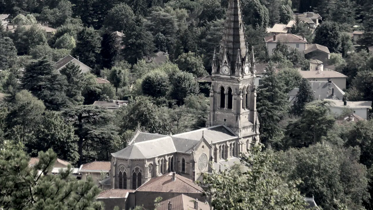 Coteau boisé des monts d'Or où des maisons dispersées s'étagent à différentes hauteurs autour du clocher néogothique d'une église de village.