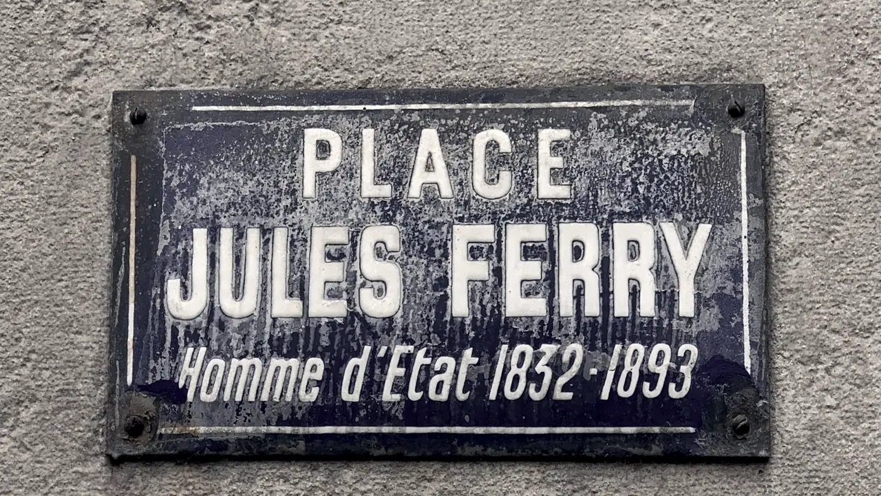 Plaque de rue émaillée vissée par ses quatre coins sur un mur crépi, avec le nom d’une place en lettres capitales et les dates d’un homme d’État.