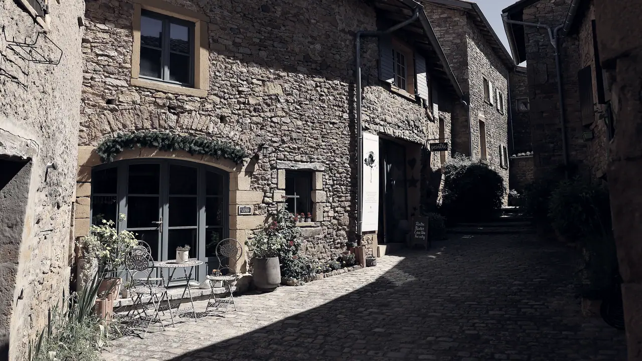 Ruelle pavée d'un village du Beaujolais bordée de maisons en pierre dorée, avec une terrasse de café à l'ombre sur la gauche.