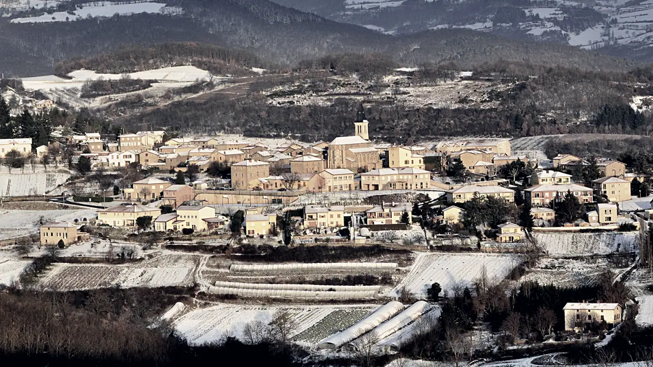Village groupé autour du clocher de son église, entouré de champs en terrasses sous la neige et de collines boisées au fond.