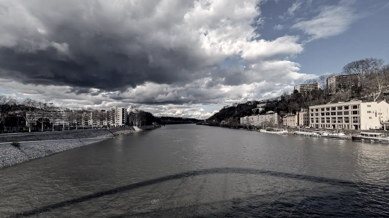 Large rivière entre deux rives bâties, avec des péniches amarrées le long du quai et un ciel chargé de nuages au-dessus de la vallée.