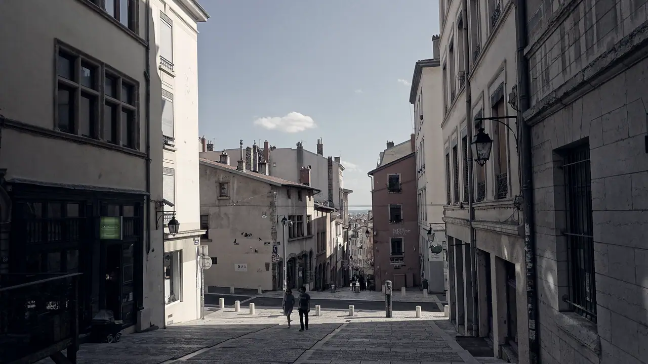 Rue pavée en pente entre de vieux immeubles, avec deux passants qui descendent vers les toits de la ville ouverte au fond.