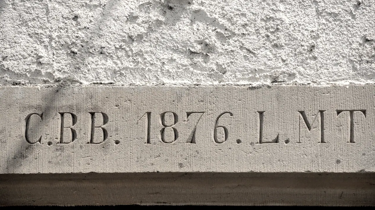 Linteau de pierre d'une porte ancienne où sont gravés le millésime 1876 et deux groupes d'initiales, sous un mur crépi éclairé par le soleil.