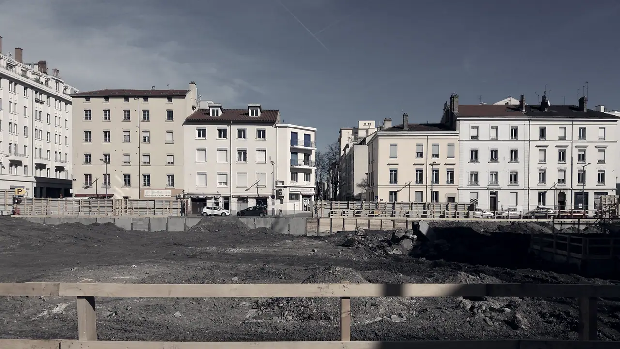Terrain excavé d'un chantier au premier plan, fermé par une barrière en bois, avec des immeubles anciens alignés derrière.