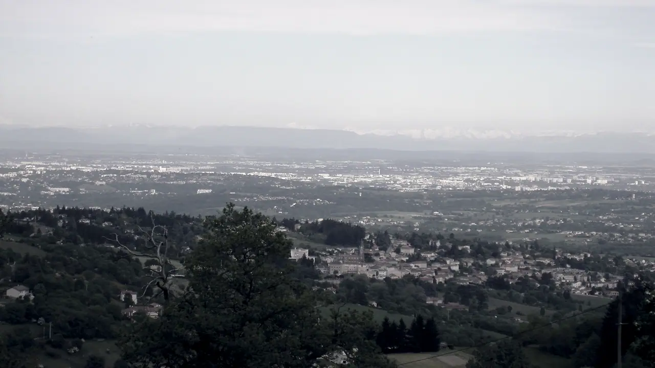 Vue plongeante depuis les collines de Vaugneray, avec le bourg dans la verdure au premier plan, la plaine urbaine de Lyon au loin et des sommets enneigés sur l'horizon.