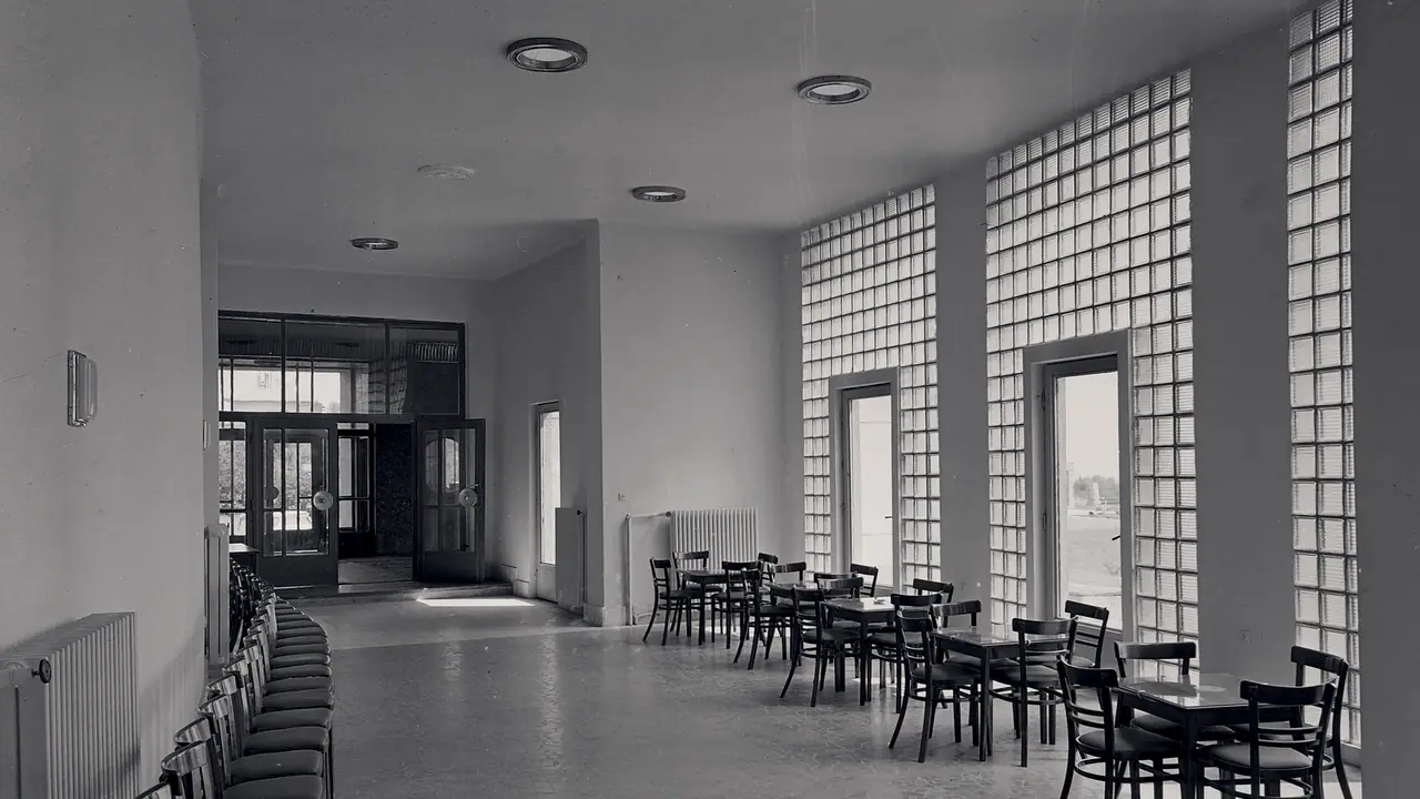 Longue salle d'attente vide : une rangée de chaises le long du mur de gauche, des tables et leurs chaises devant les hautes parois de briques de verre à droite.