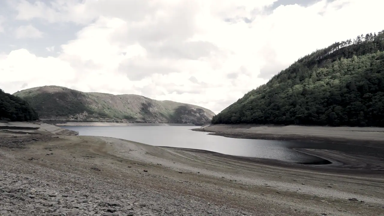 Lac de barrage presque vidé entre deux collines boisées : l'eau ne remplit plus que le fond de la vallée, au milieu de larges rives de galets mises à nu.