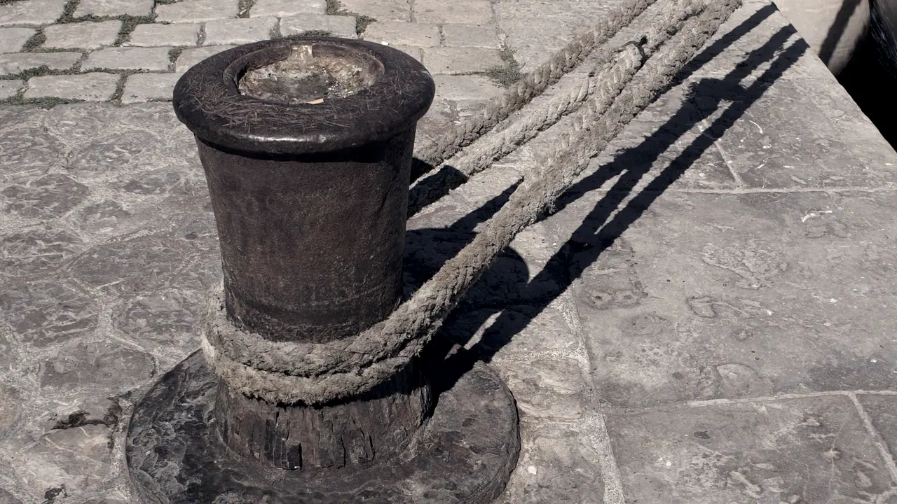 Vieille bitte d'amarrage noircie et rongée par le temps, sur un quai dallé, avec un cordage de chanvre tourné deux fois à sa base et des amarres tendues vers le bord du quai.
