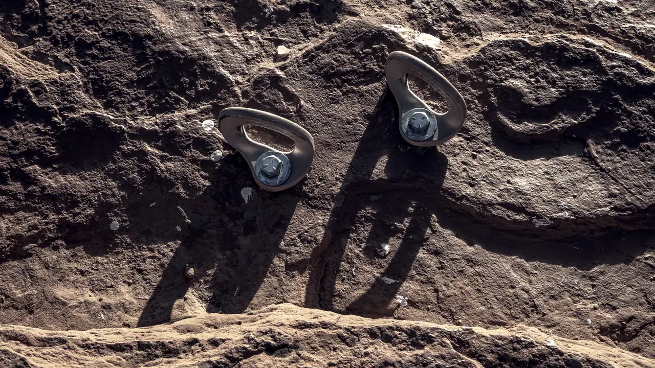Deux plaquettes d'ancrage d'escalade vissées côte à côte dans un grès sculpté par l'érosion, chacune tenue par son boulon, avec leurs ombres portées sur la roche.