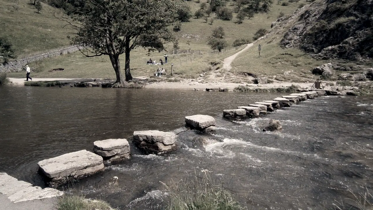 Pierres de gué alignées en travers d'une rivière peu profonde, depuis la rive du premier plan jusqu'au sentier qui remonte sur l'autre berge.