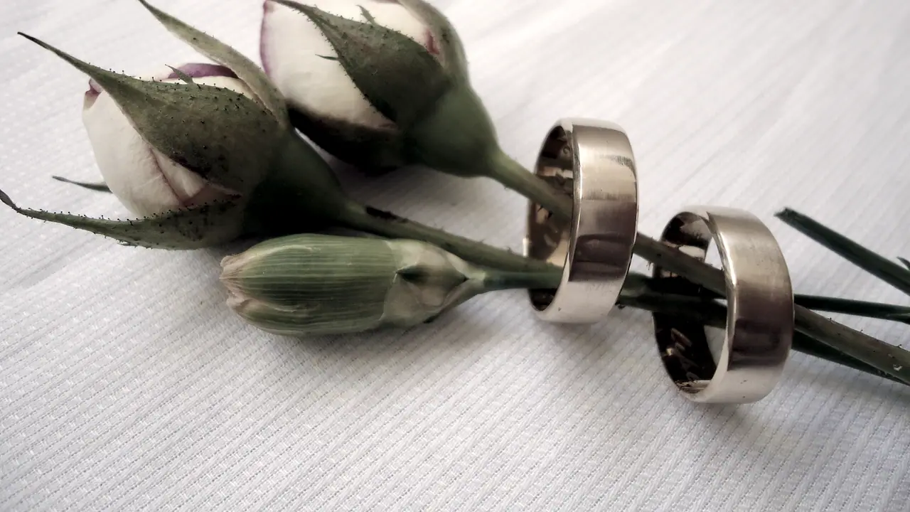 Deux alliances larges enfilées sur les tiges de trois fleurs encore en bouton, posées sur une étoffe tissée.
