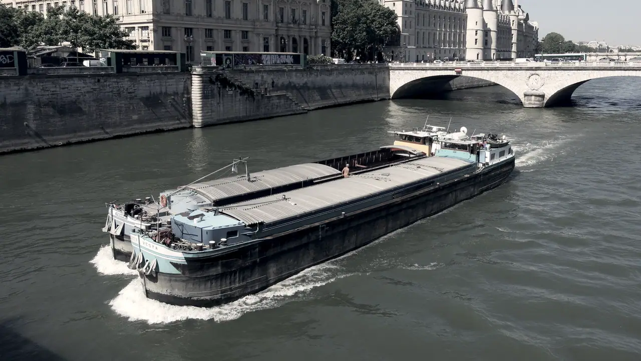 Deux péniches amarrées à couple qui naviguent ensemble sur un fleuve, coque contre coque, devant un quai de pierre et ses façades anciennes.
