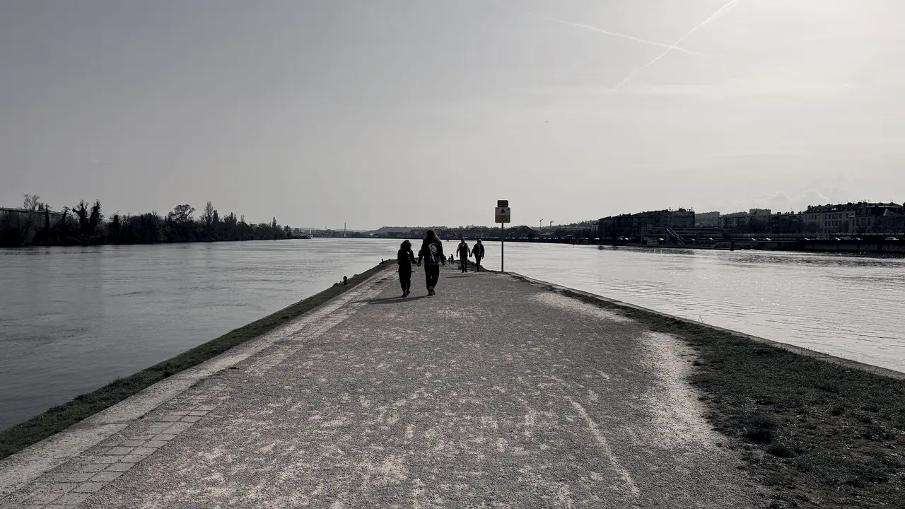 Pointe de terre gravillonnée entre deux cours d'eau qui se rejoignent, avec des promeneurs sur le chemin et la ville au fond.