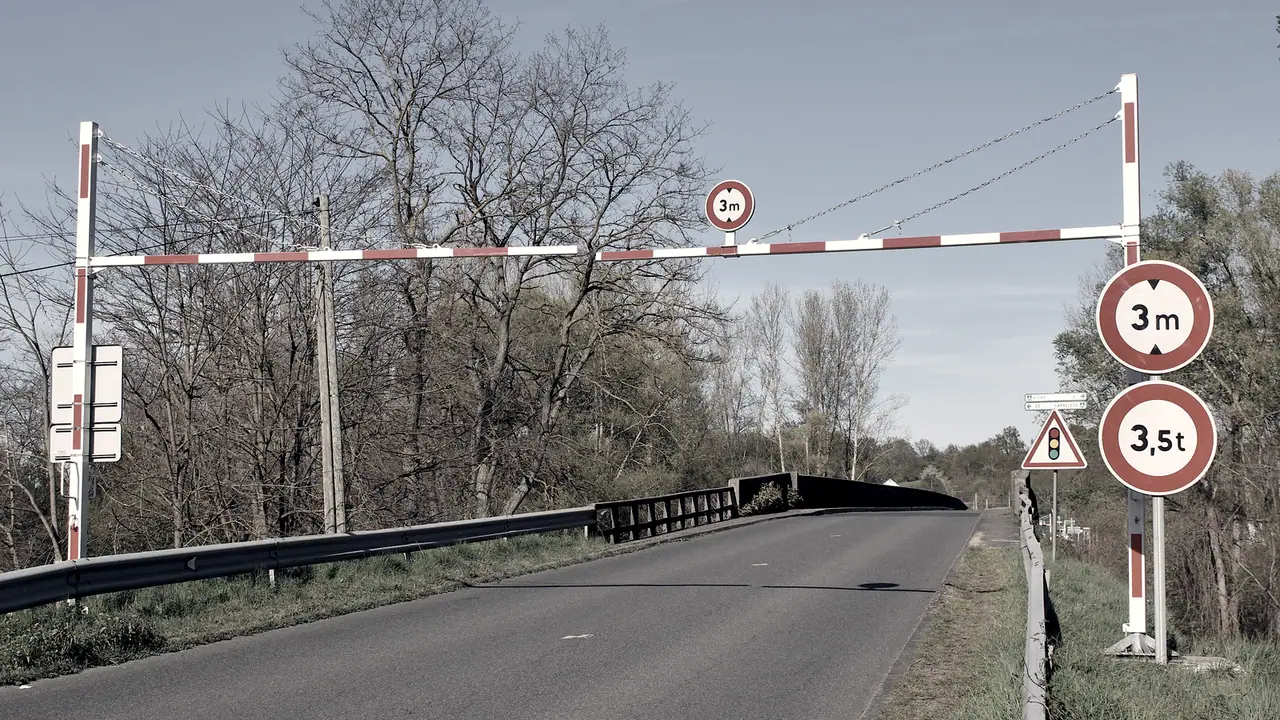 Portique de limitation fixé au-dessus d'une route, sa barre horizontale rouge et blanche barrant l'accès à un pont étroit.