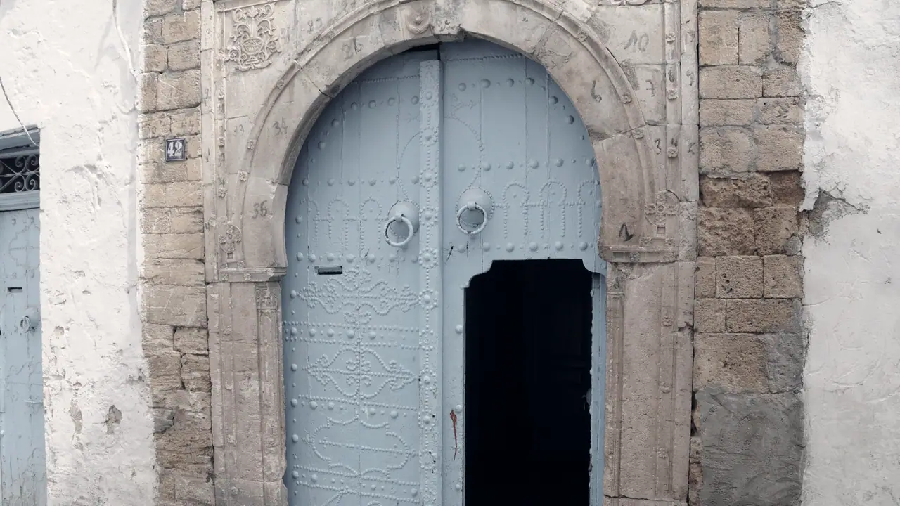 Grande porte cloutée à deux battants sous un arc de pierre sculpté, dont seule la petite porte découpée dans un battant est ouverte sur l'ombre.