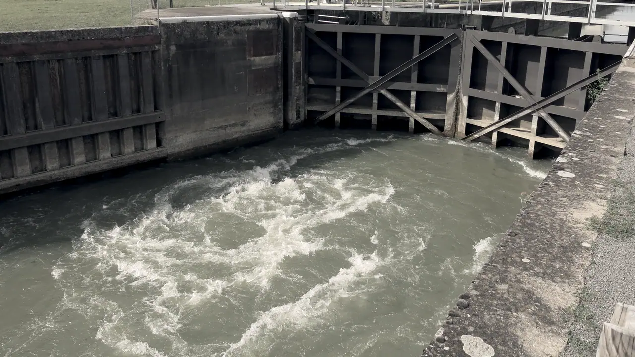 Sas d'une écluse en train de se vider : l'eau bouillonne au pied des portes fermées, renforcées de croisillons de métal, entre deux murs de béton.