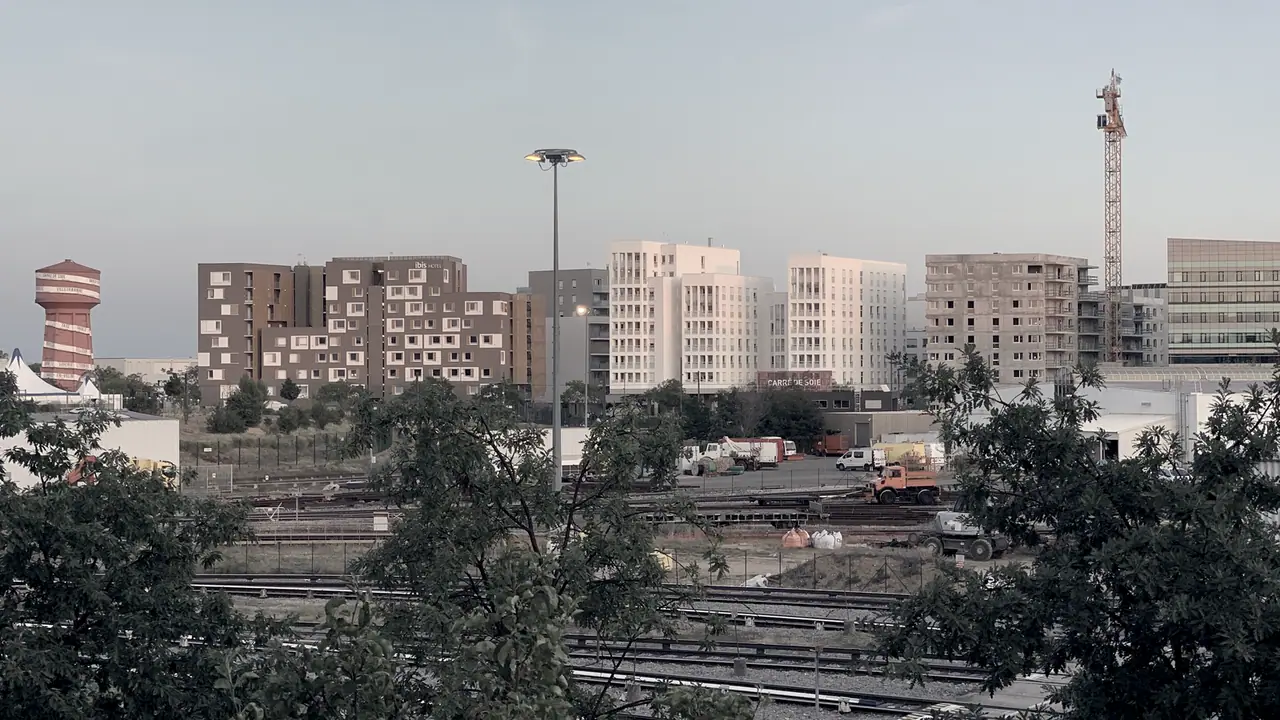 Rangée d'immeubles récents alignés derrière des voies ferrées, avec une grue de chantier au fond et des arbres au premier plan.