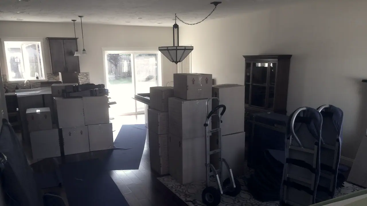 Pièce de vie encombrée de cartons de déménagement empilés, avec deux diables et un meuble poussé contre le mur.