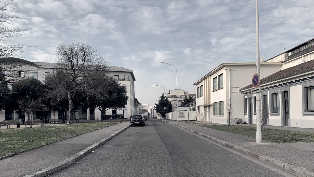 Rue déserte bordée d'arbres nus et de bâtiments bas, avec une voiture stationnée au loin sous un ciel nuageux.