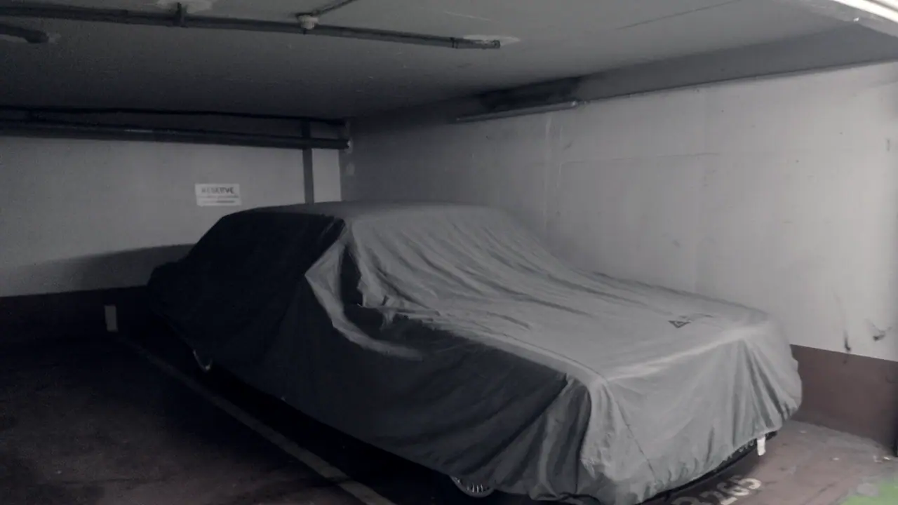 Voiture entièrement couverte d'une housse grise, garée sur sa place dans un parking souterrain, sous les tuyaux qui courent au plafond.