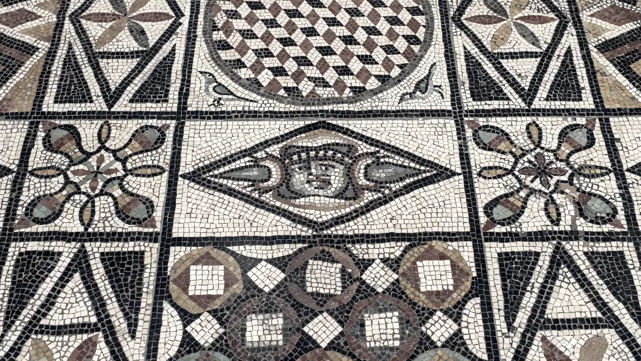 Pavement de mosaïque romaine divisé en panneaux, chacun orné d'un motif différent : losanges, rosaces, damier, et un masque au centre.