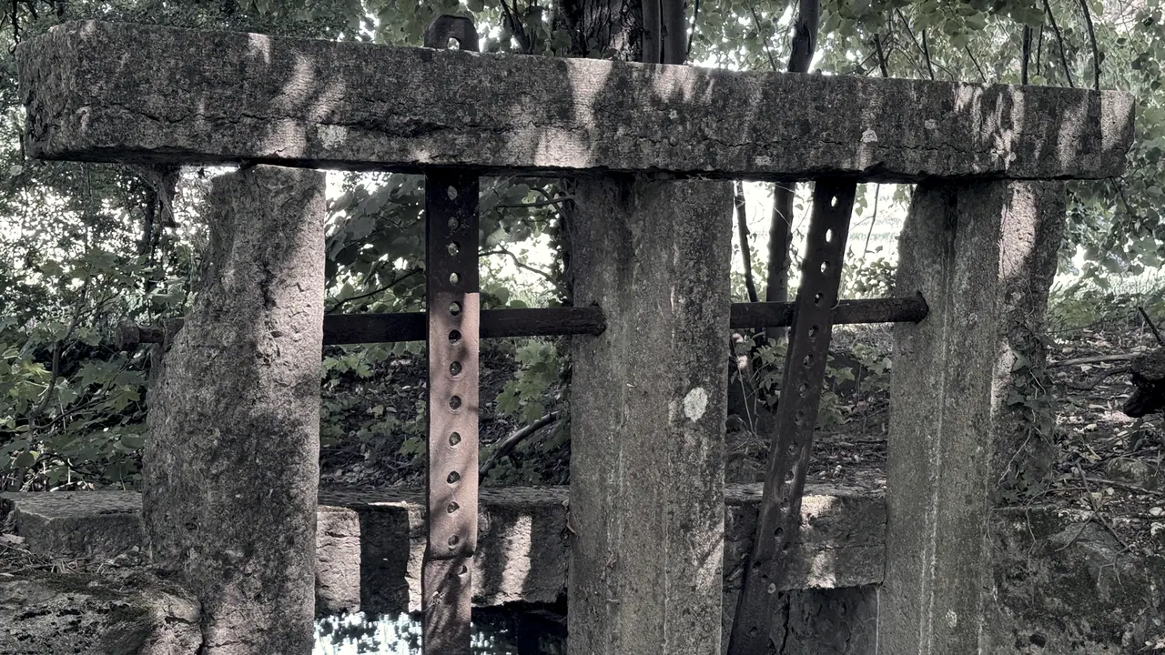 Ancien seuil de rivière : entre deux piles de pierre moussues, une barre de fer percée de trous et des traverses de bois forment la vanne.