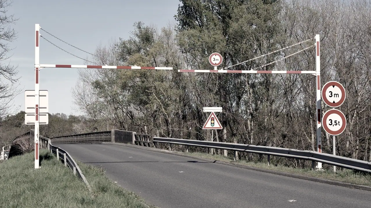 Portique de limitation de hauteur rayé de rouge et de blanc au-dessus d'une route, à l'entrée d'un pont, avec les panneaux de trois mètres et de 3,5 tonnes.