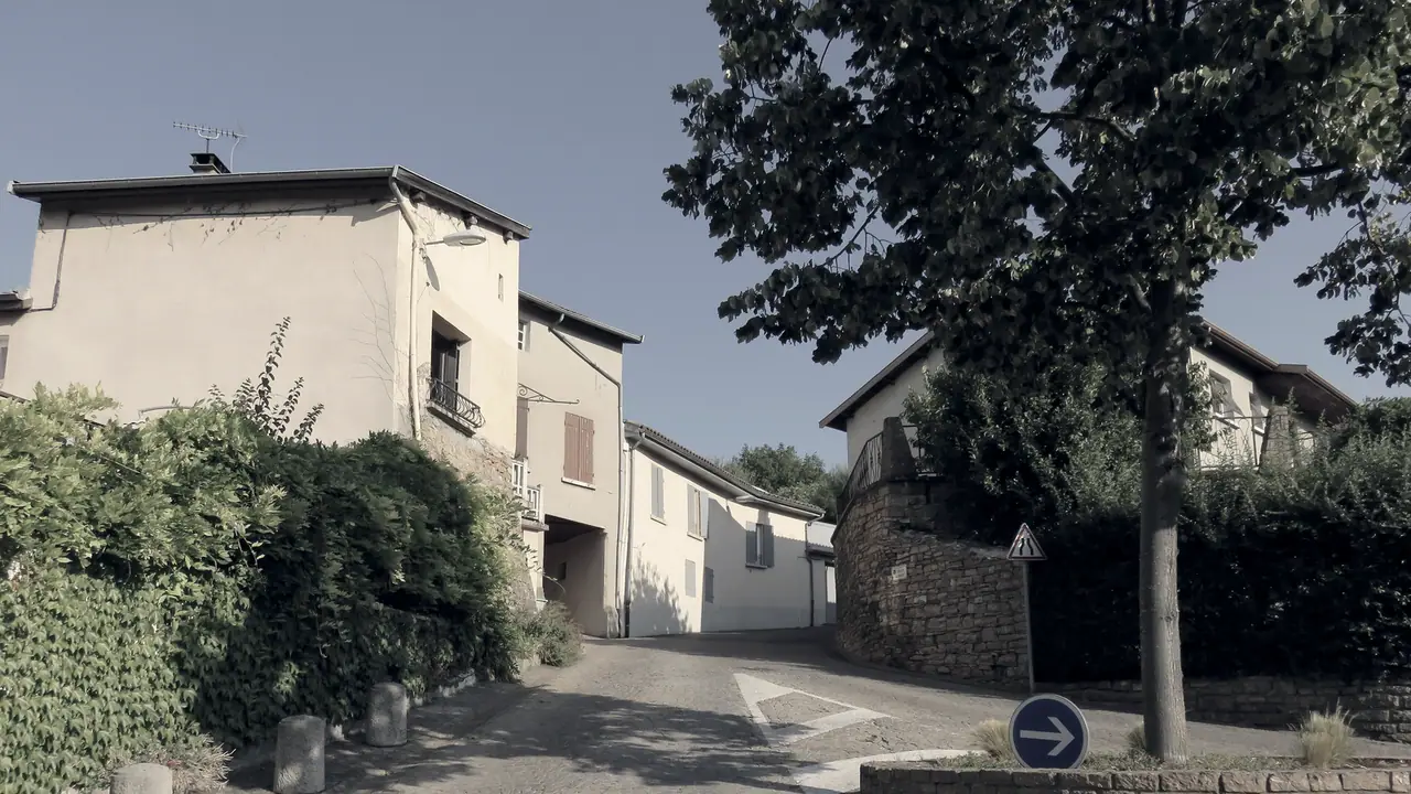 Rue pavée d'un village en pente, bordée de maisons crépies aux volets clos, d'un muret de pierre dorée et d'un arbre planté sur un rond-point.