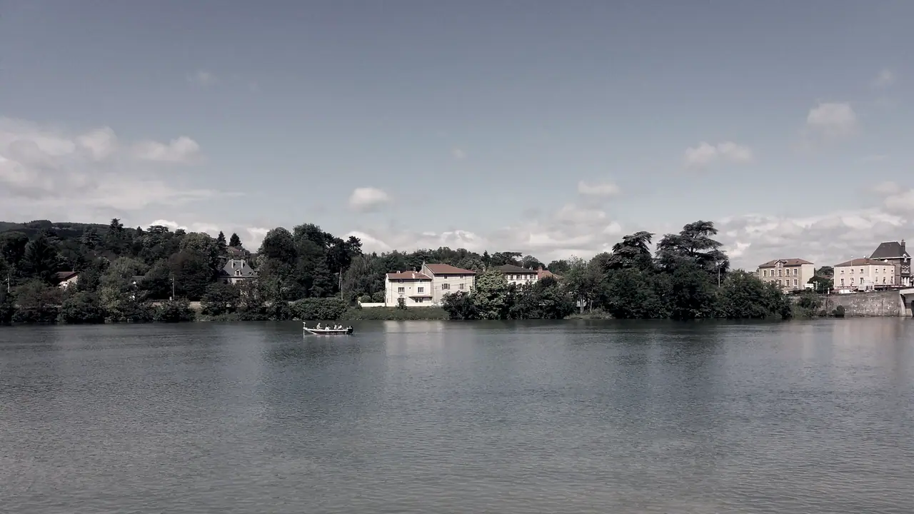 Rive boisée de la Saône vue depuis un quai, avec des maisons aux toits de tuiles entre les arbres et une petite barque sur l'eau calme.