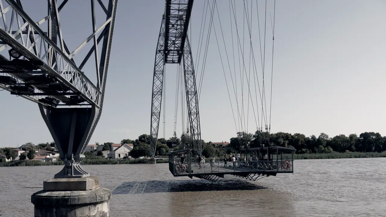 Nacelle d'un pont transbordeur suspendue à ses câbles au-dessus d'une rivière, entre le pied d'un pylône métallique et une rive bordée de maisons.