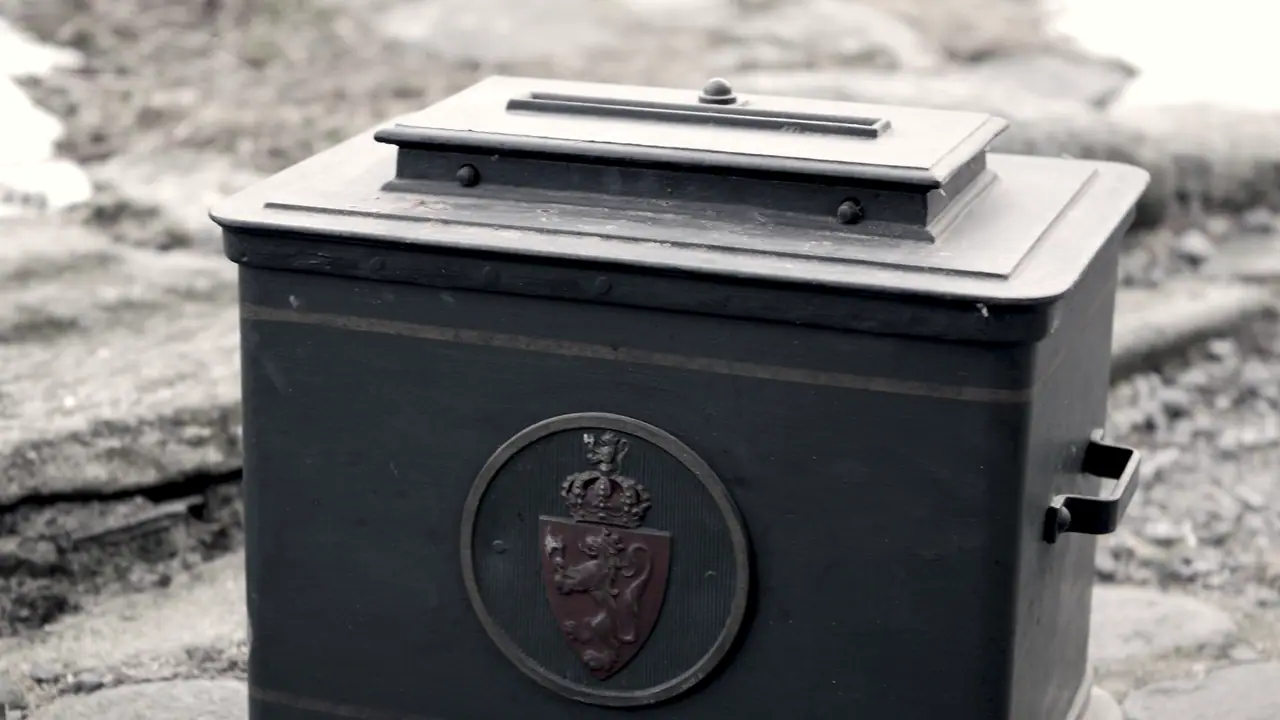 Urne de vote ancienne en métal sombre, au couvercle percé d'une fente, frappée d'un écusson au lion couronné et posée sur des pierres plates.