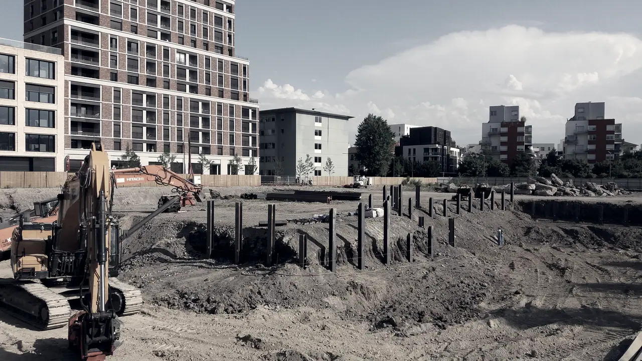 Pelleteuse à chenilles devant une fouille de chantier hérissée de pieux, avec des immeubles récents à l'arrière-plan.
